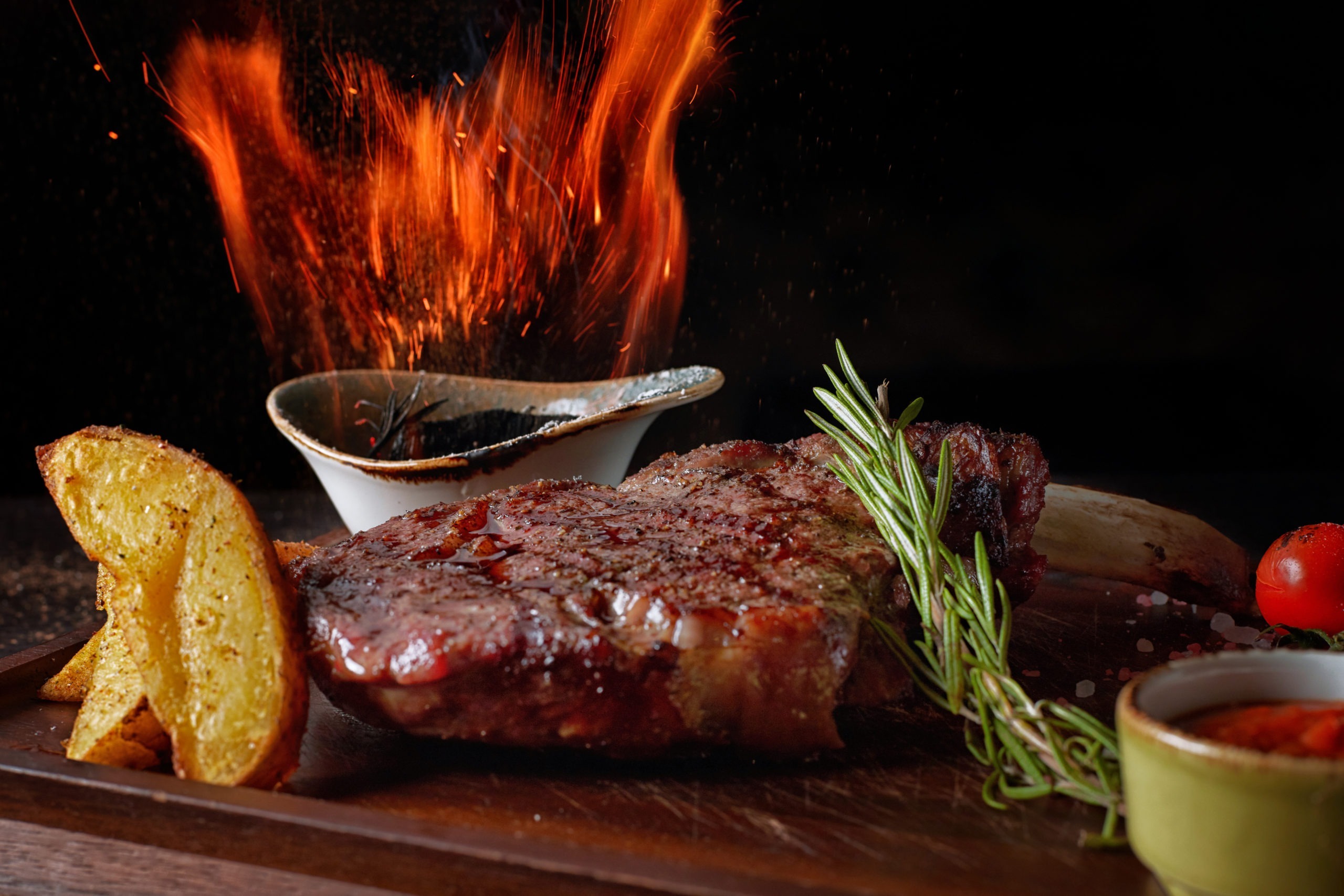 Meat steak with fire, on a wooden board, with potatoes and sauce, on a black background